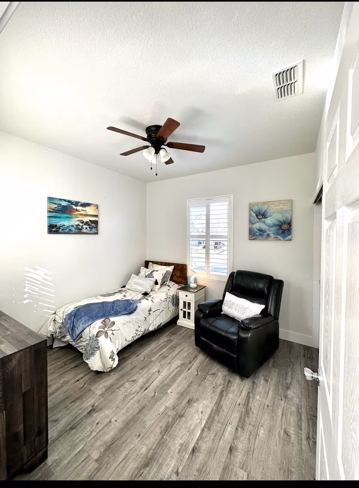 A cozy bedroom with a twin bed, a black recliner, nightstand, ceiling fan, and art.