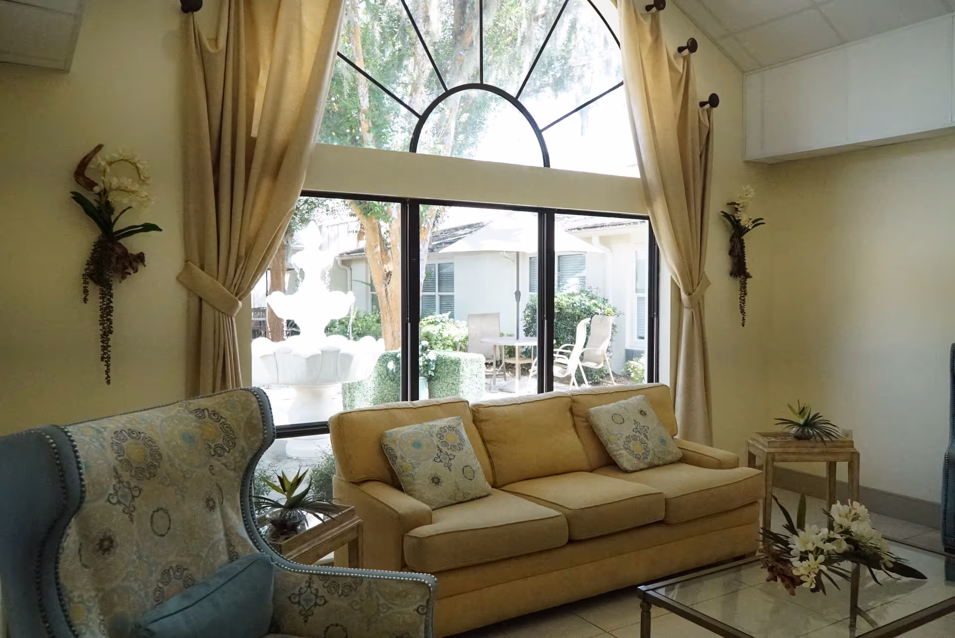 Bright living room with beige sofa, blue chair, glass coffee table, and arched window with curtains.