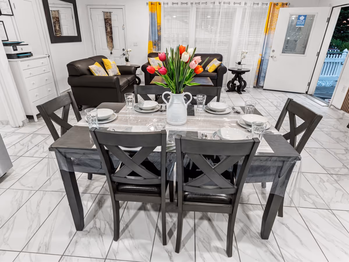 Six-seat dining table set with plates, glasses, and a tulip centerpiece in a bright common area.
