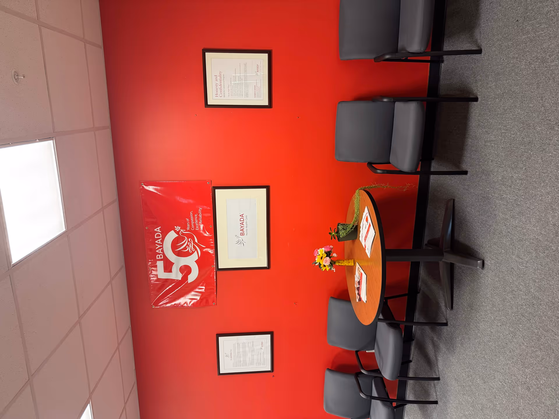 Interior waiting area with a red wall, framed certificates, BAYADA banner, and round table with chairs.