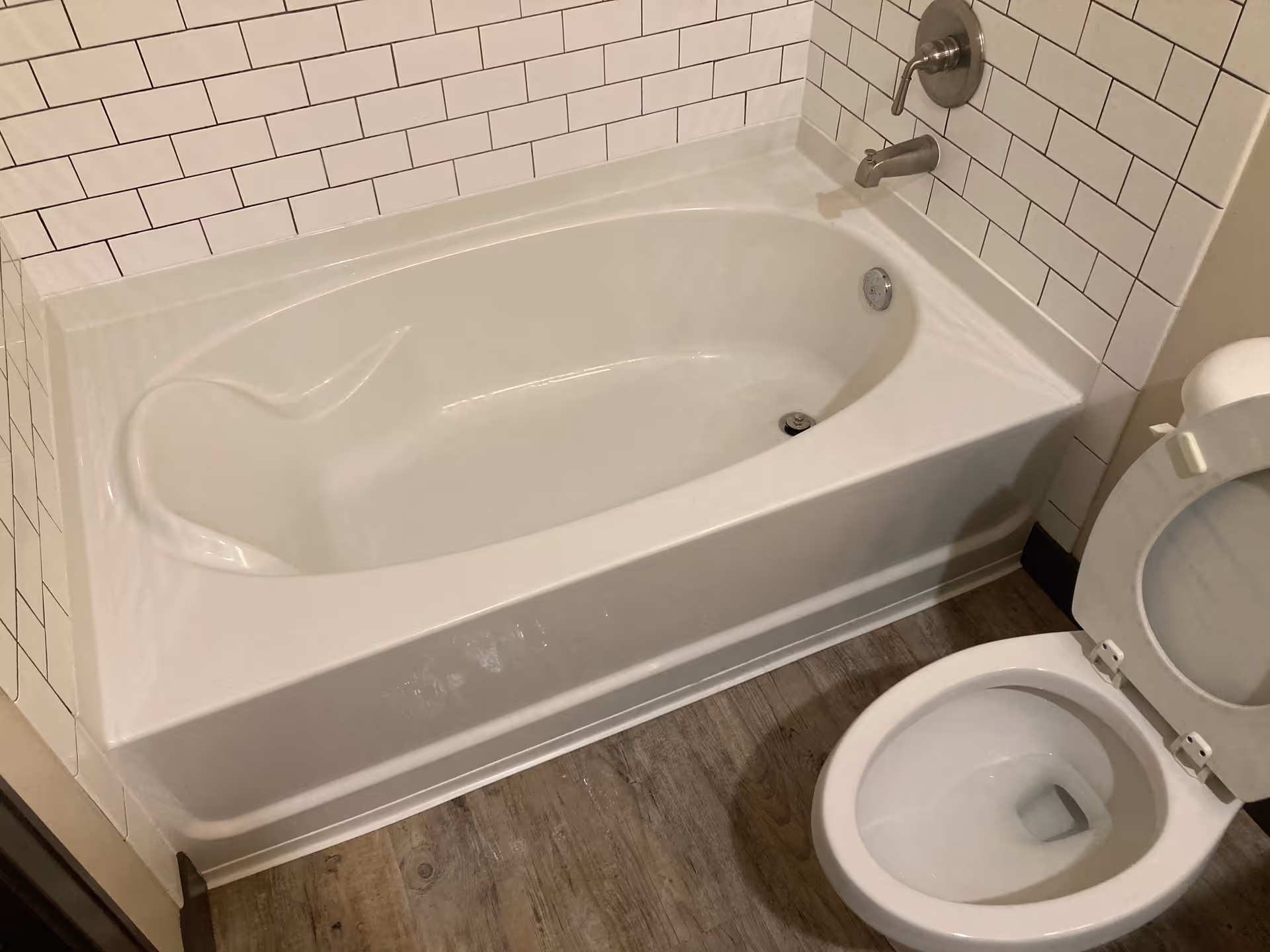 White bathtub and toilet in a tiled bathroom with wood-look flooring.