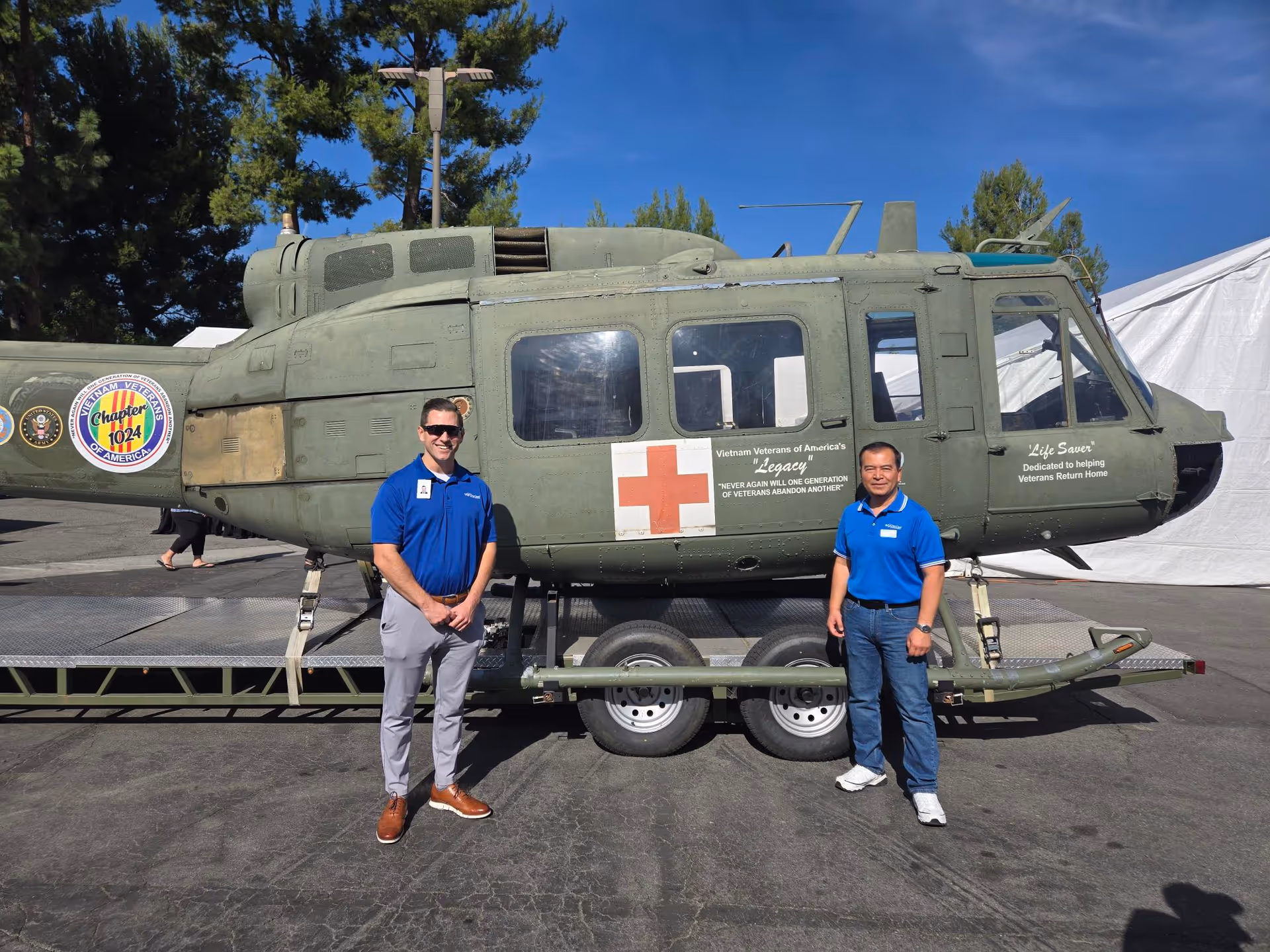 Two men in blue shirts stand beside a military helicopter on display outdoors.