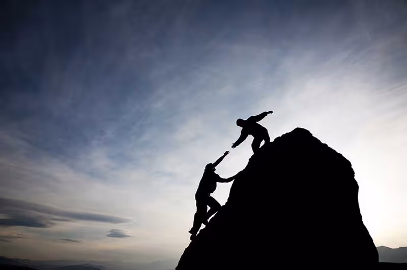 Two silhouetted climbers help each other ascend a rocky peak at sunset.