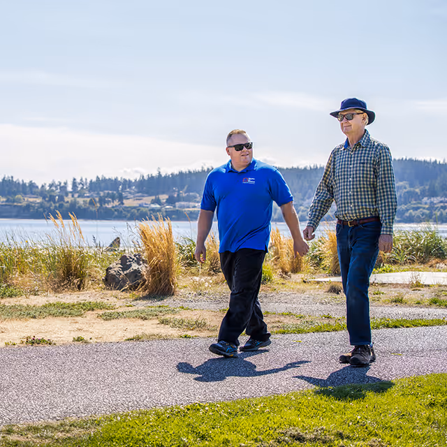 Two men walk hand-in-hand along a waterfront path with grasses and hills behind.