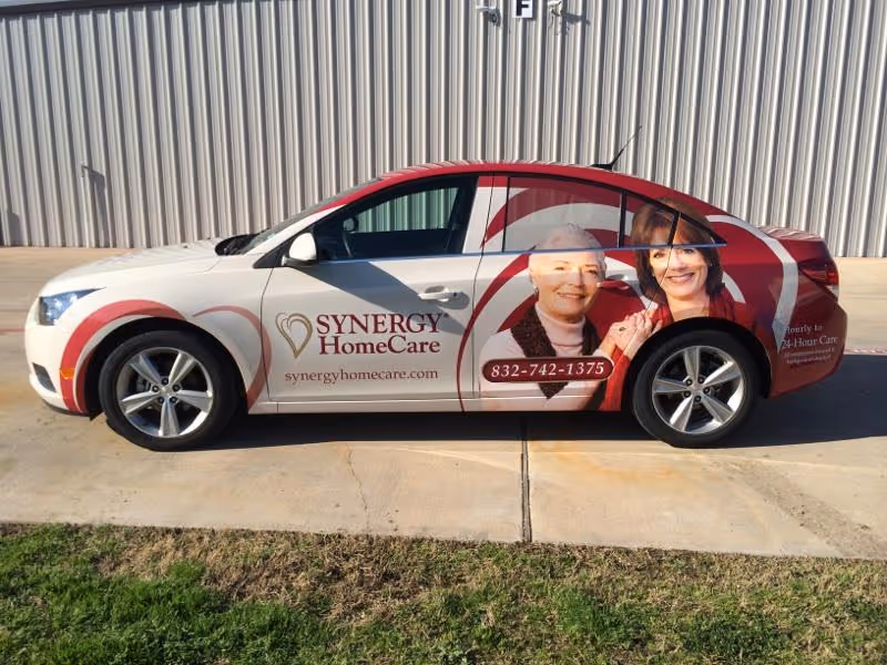 Synergy HomeCare branded car parked beside a corrugated metal building with caregiver images.