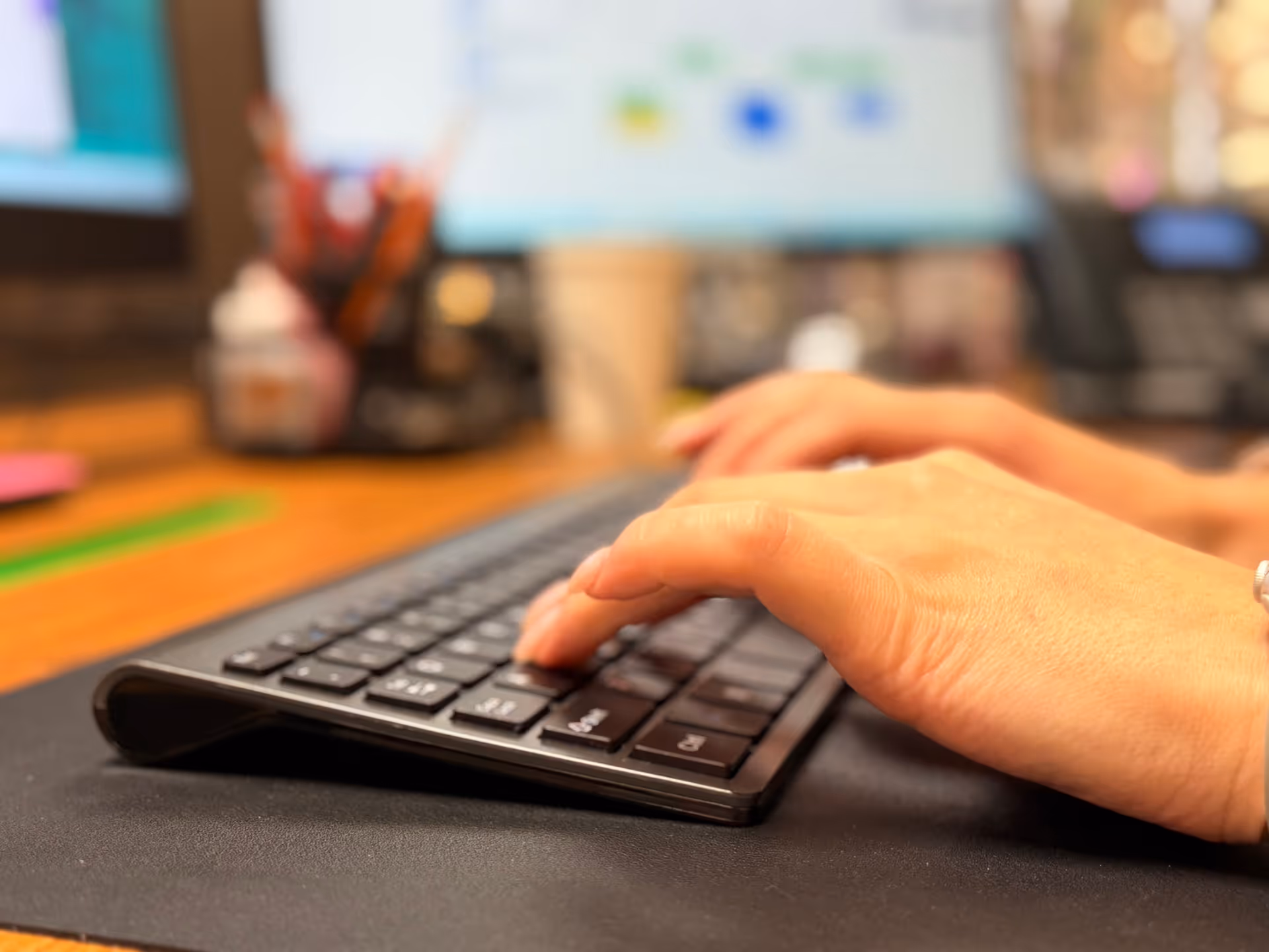 Close-up of hands typing on a keyboard at a desk with a blurred computer screen.