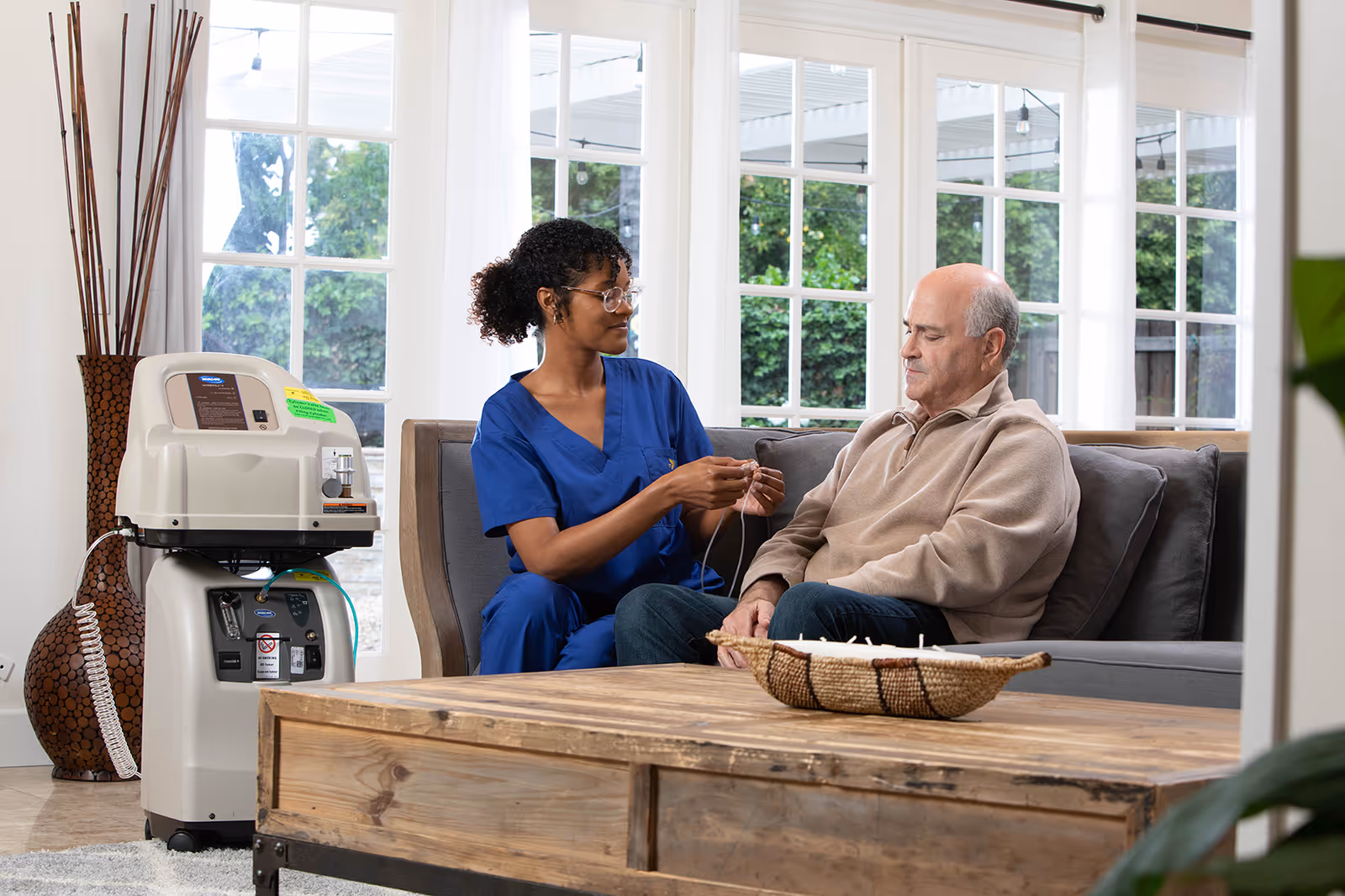 Caregiver in blue scrubs sits with elderly man on a living room couch, medical device nearby.