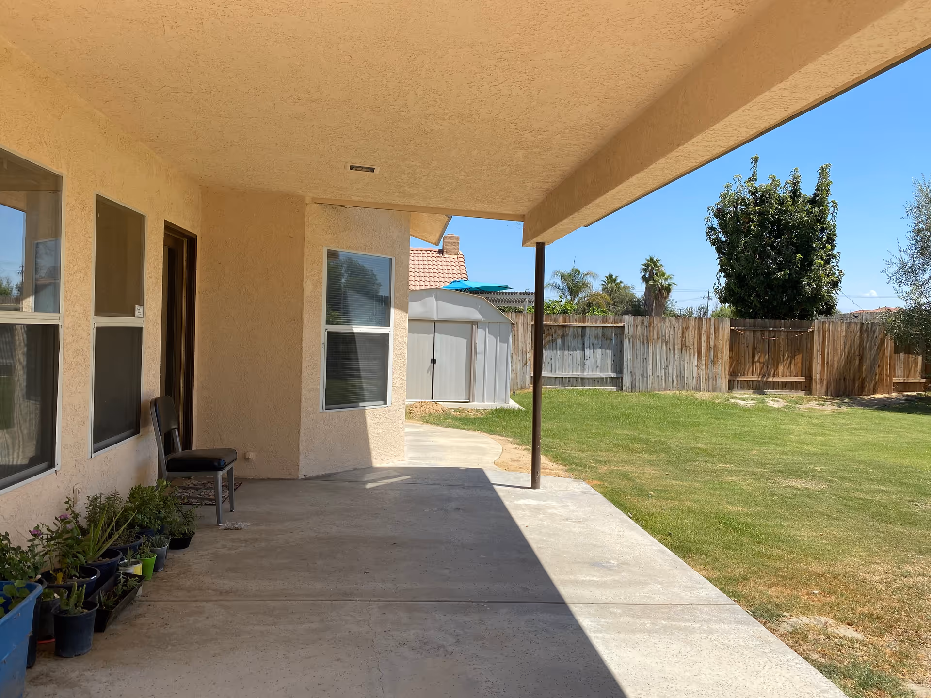 Covered patio of a beige residential building with chairs, potted plants, and a grassy yard.