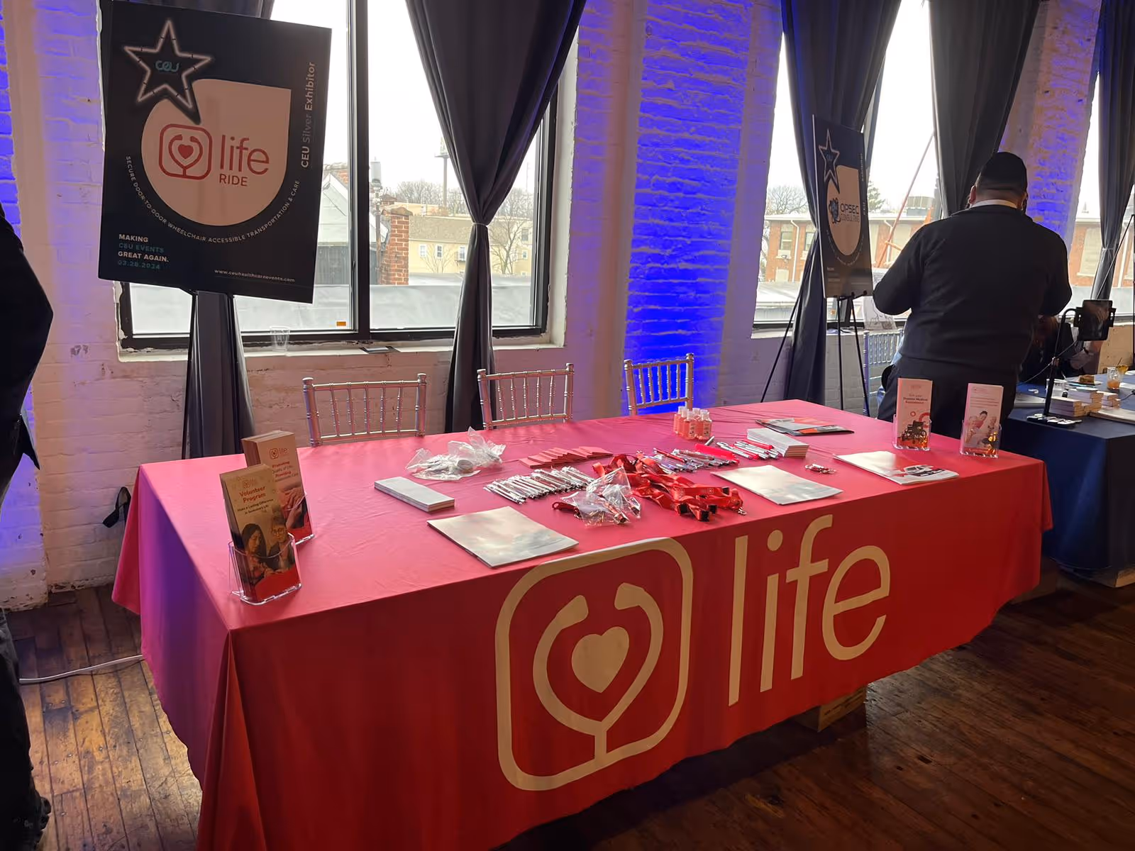 Indoor event setup with a pink Life table, banners, and attendees near windows.