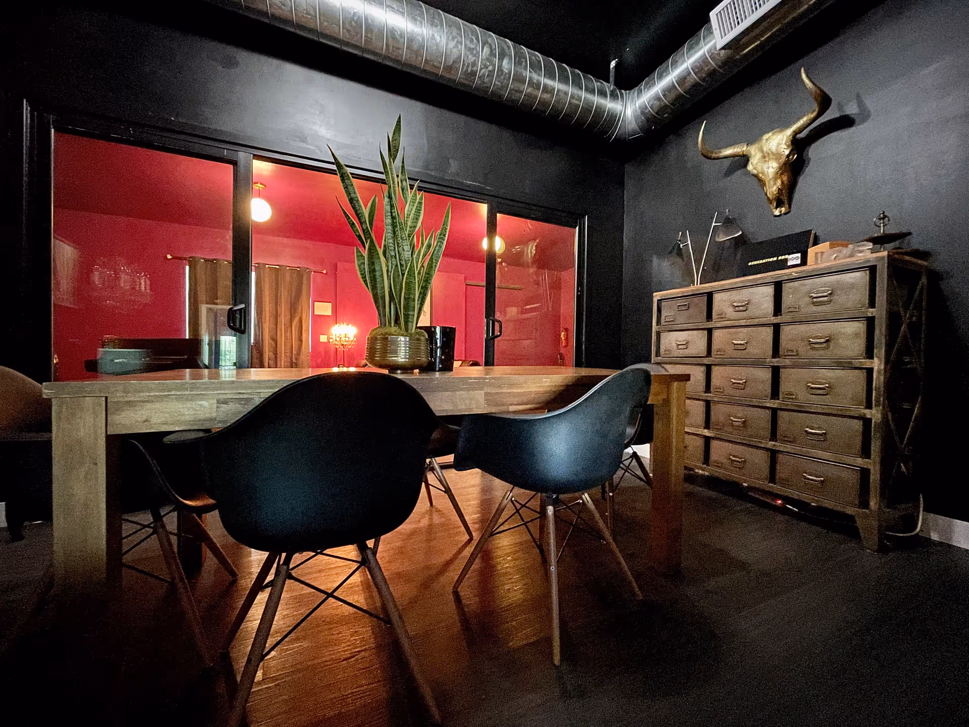 Dark interior dining area with a wooden table, black chairs, plant, and a tall cabinet.