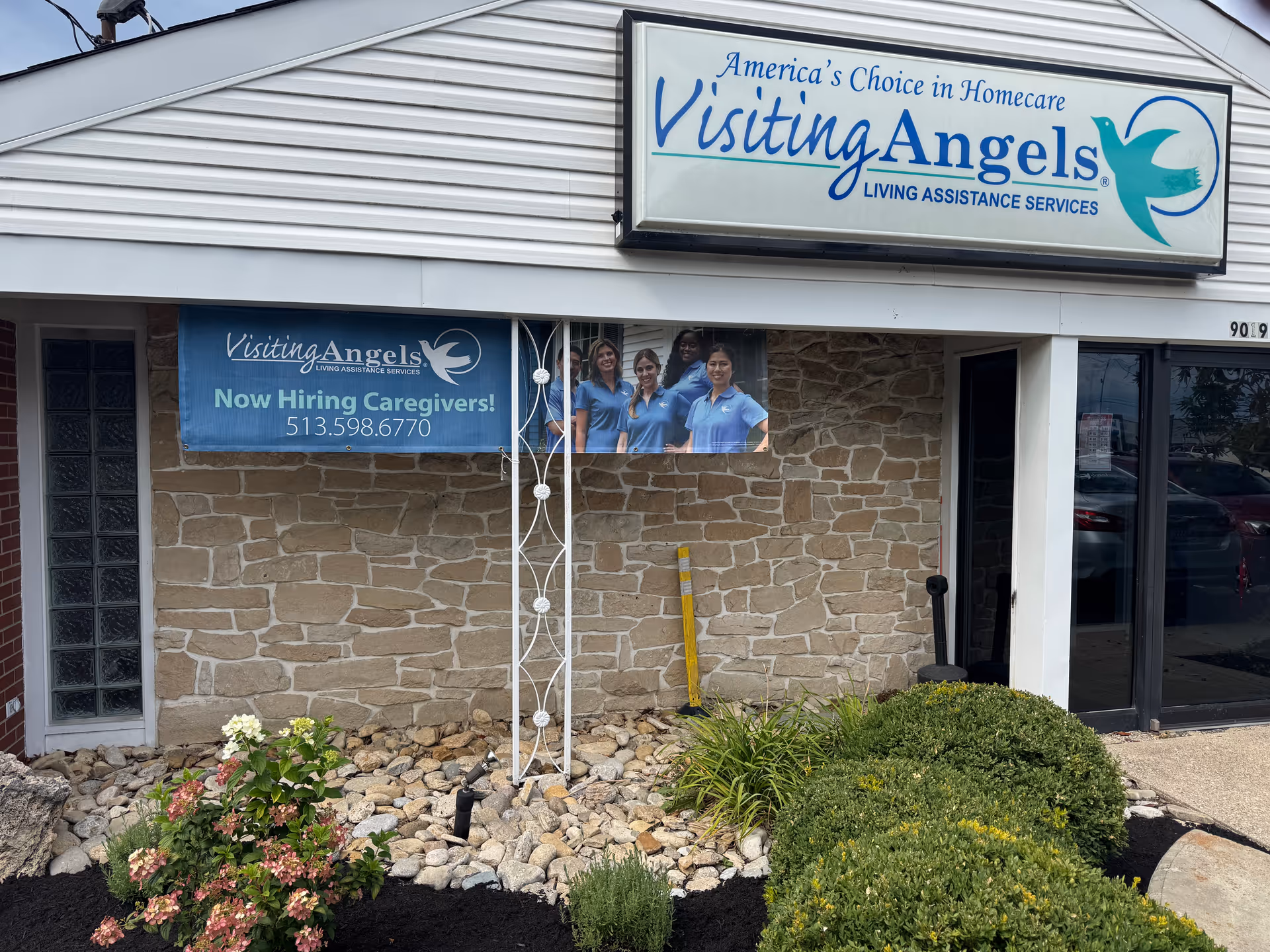 Exterior of Visiting Angels building with large sign, banners, stone wall, and landscaped entrance.