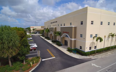 Exterior view of a beige senior living facility with a parking lot and palm trees.