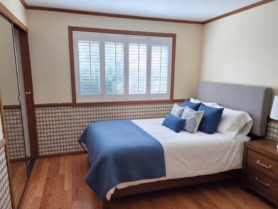 Cozy bedroom with a queen bed, blue bedding, window shutters, and plaid wainscoting.