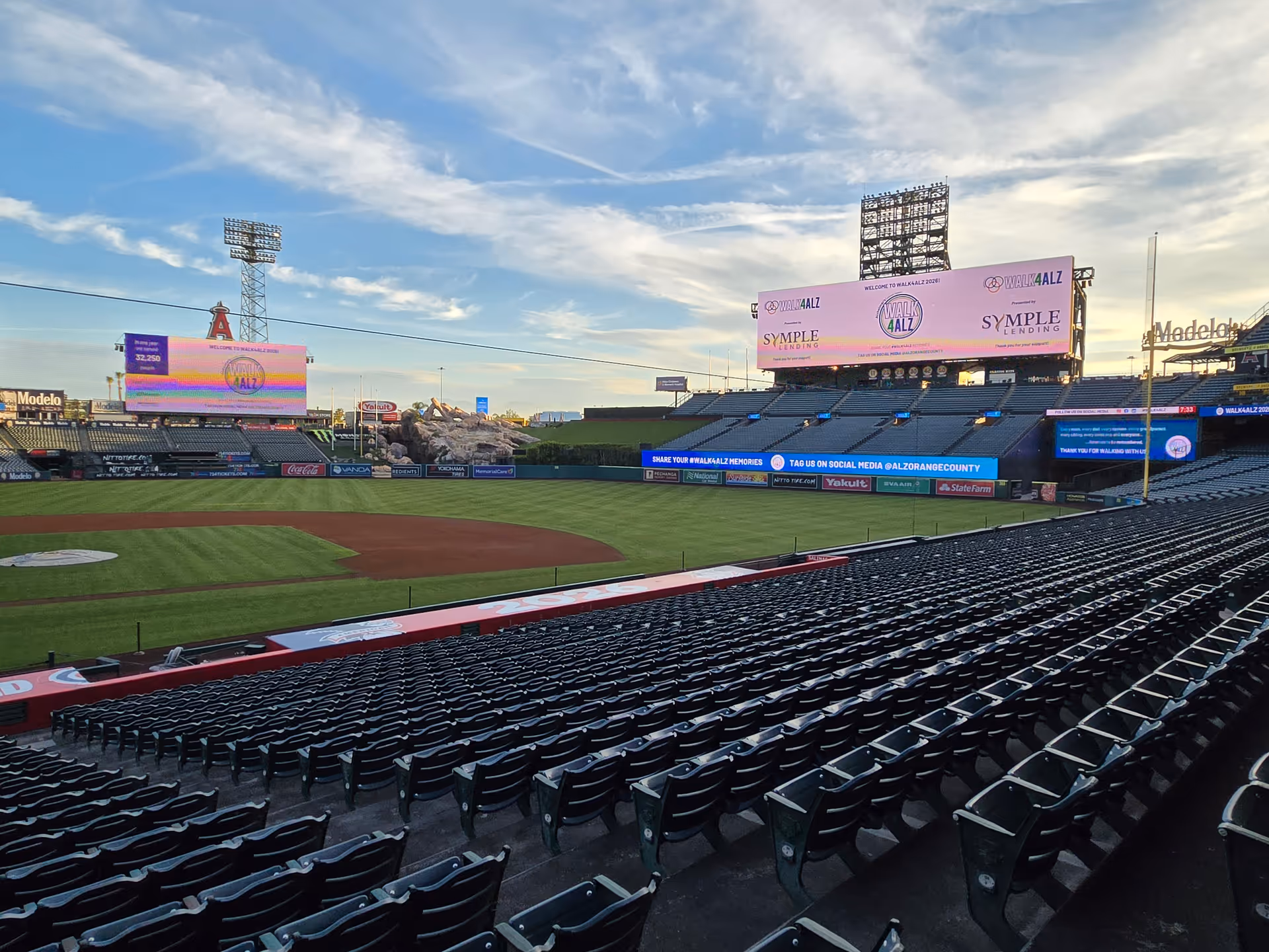 Empty baseball stadium with green field, rows of seats, and large digital scoreboards against sky.