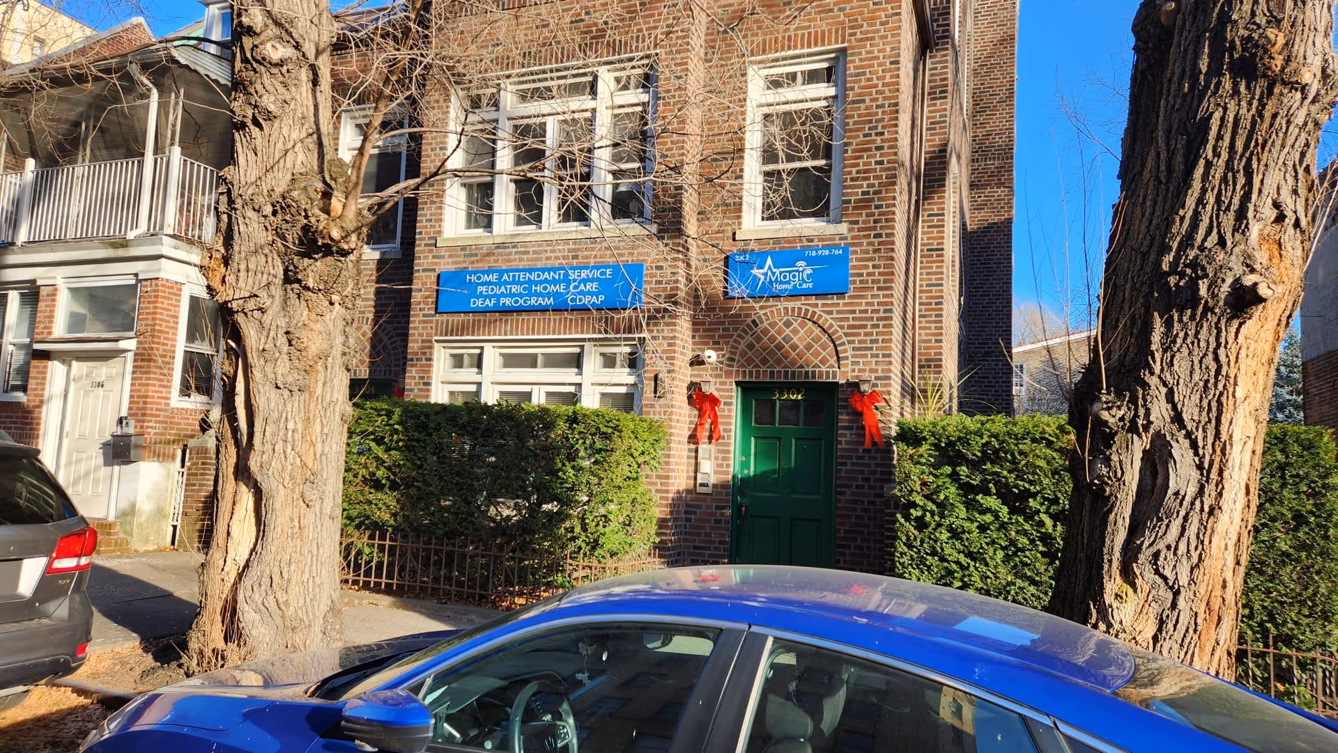 Brick building facade with blue signage, green door, red bows, and parked cars outside.