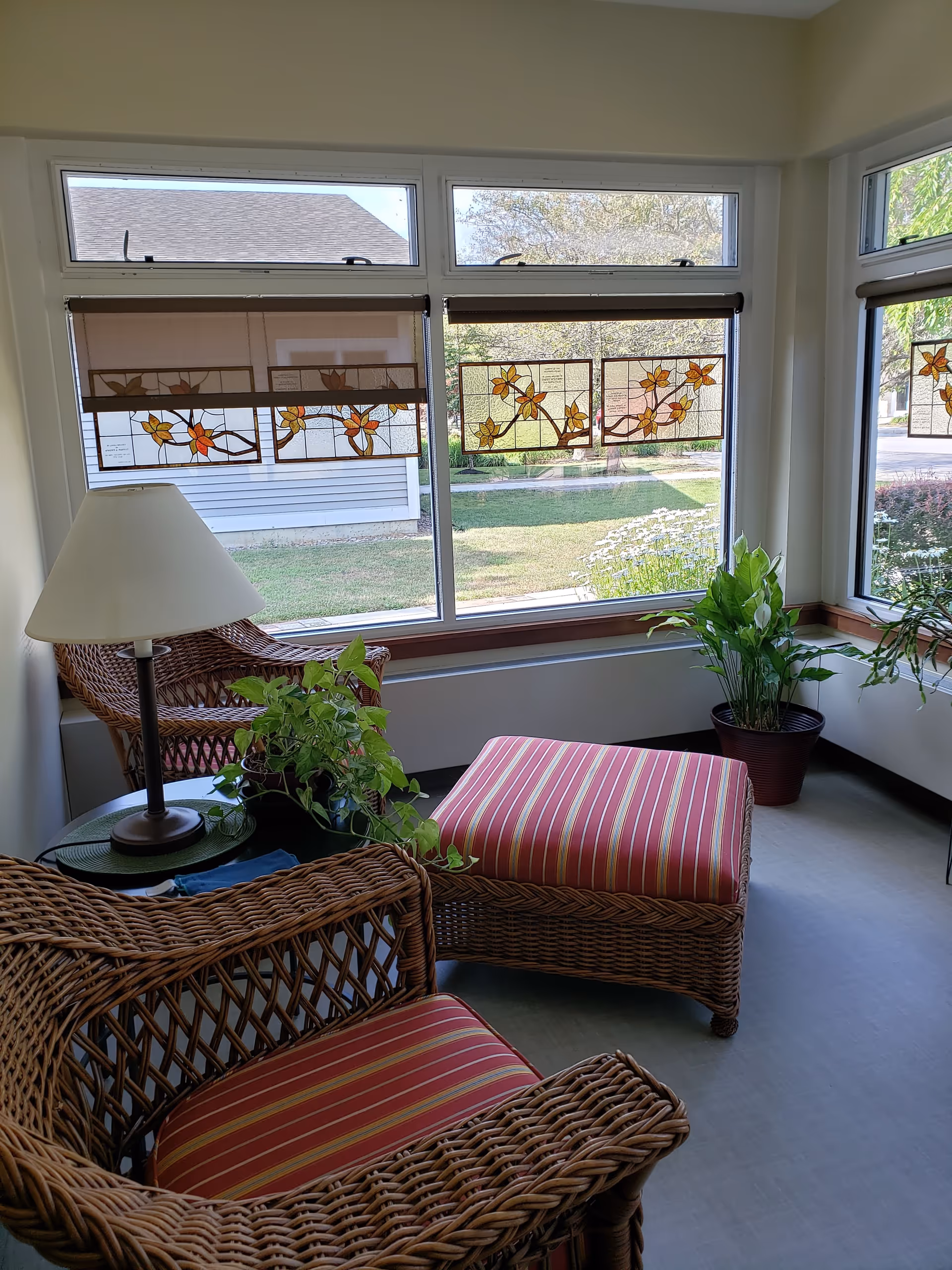 Sunlit lounge with wicker chairs, striped cushions, plants, and decorative stained glass panels.