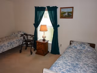 Two single beds with blue floral quilts, a nightstand, lamp, and walker in bedroom.