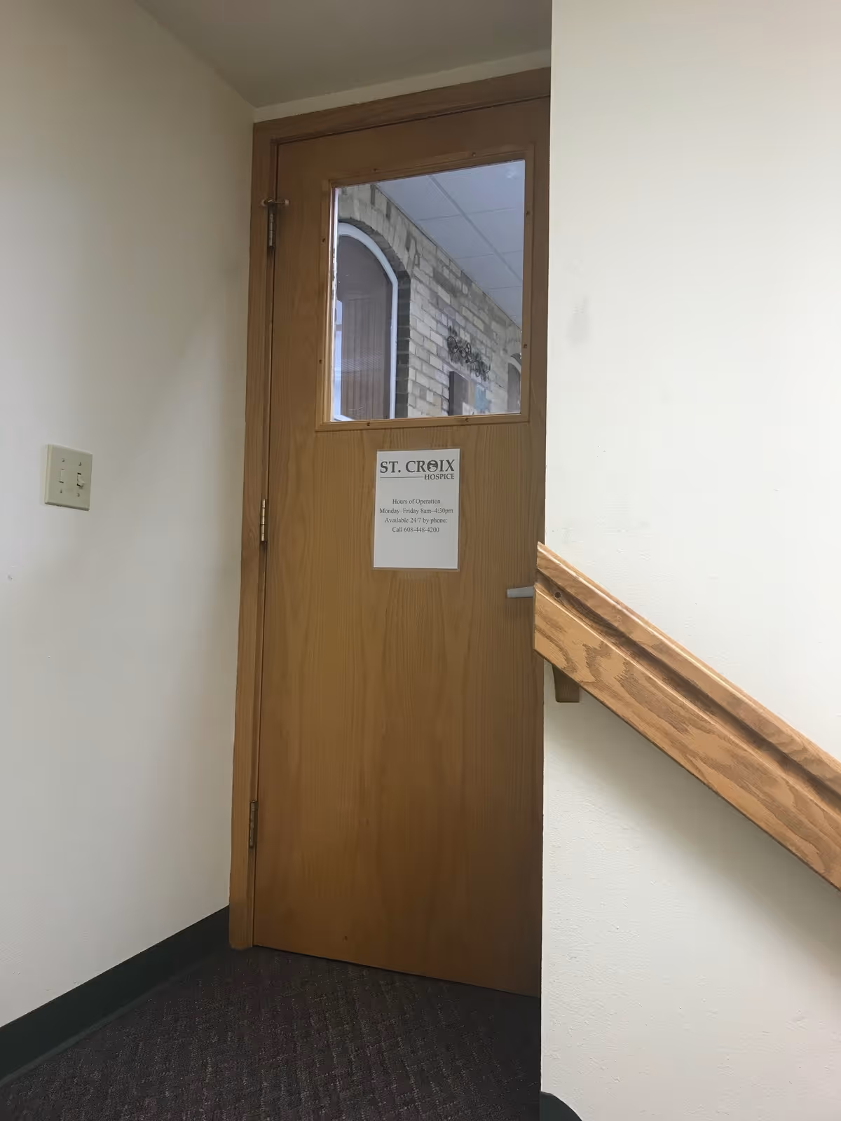 Wooden door with window in a hallway, St. Croix Hospice sign, stair railing nearby.