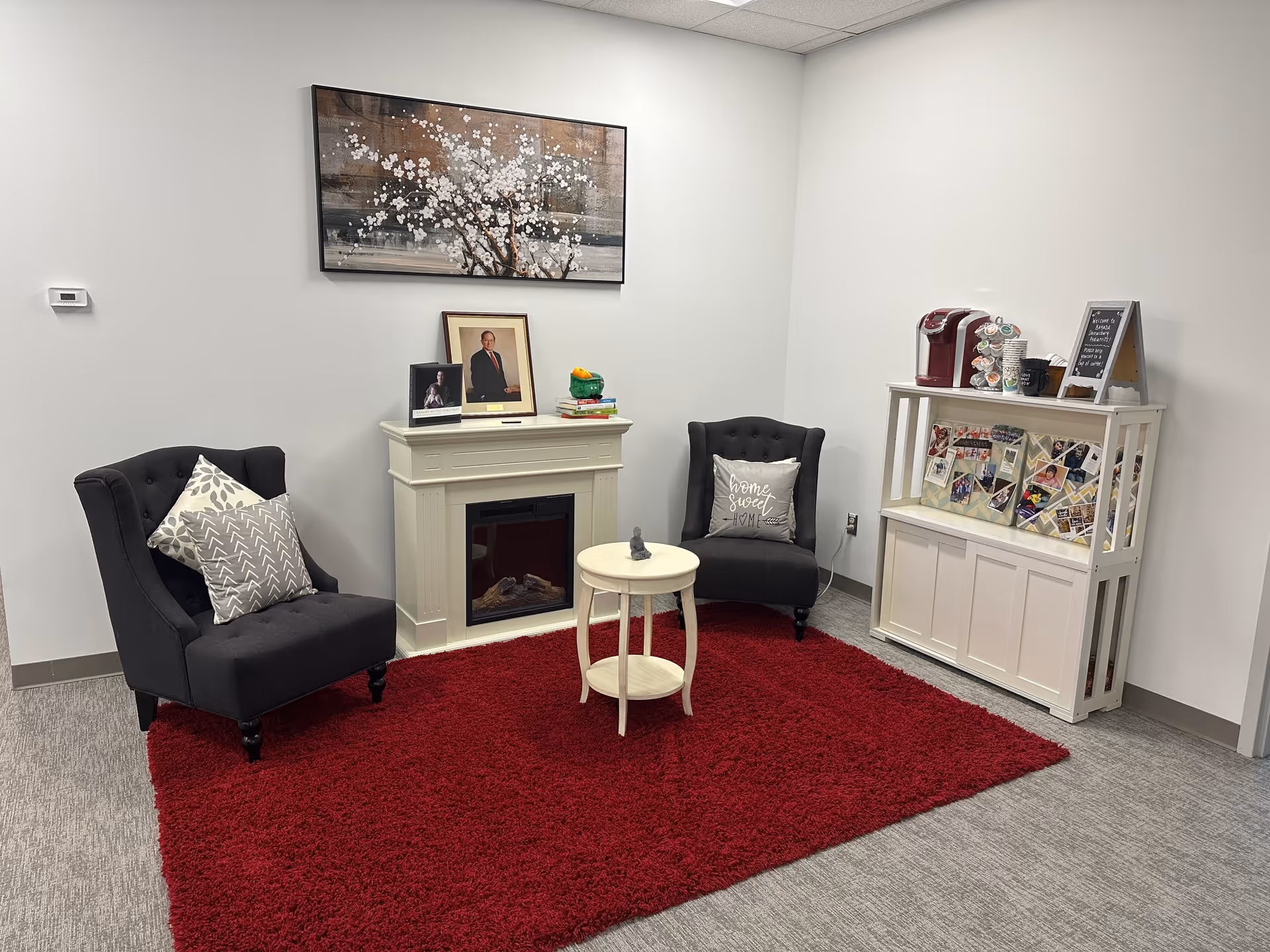 A cozy waiting lounge with two dark chairs, a white mantel, and a red rug.