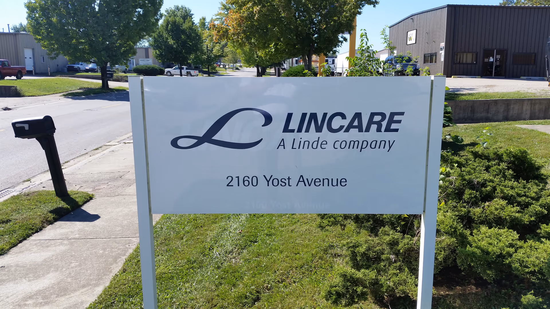A LINCARE sign on a grassy area outside a business with trees and industrial buildings behind.