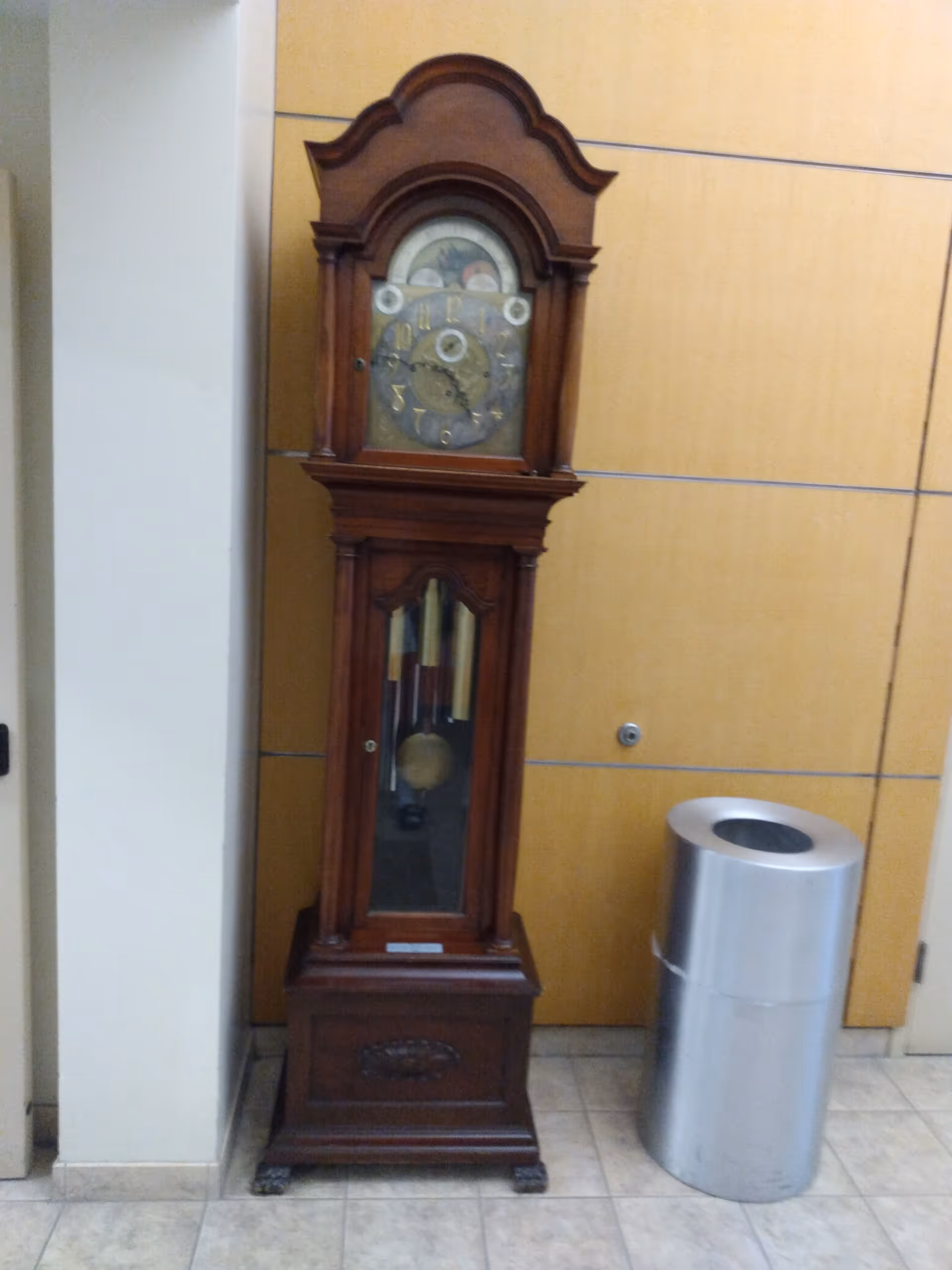 Tall wooden grandfather clock beside a metal trash can in a hall with beige walls.