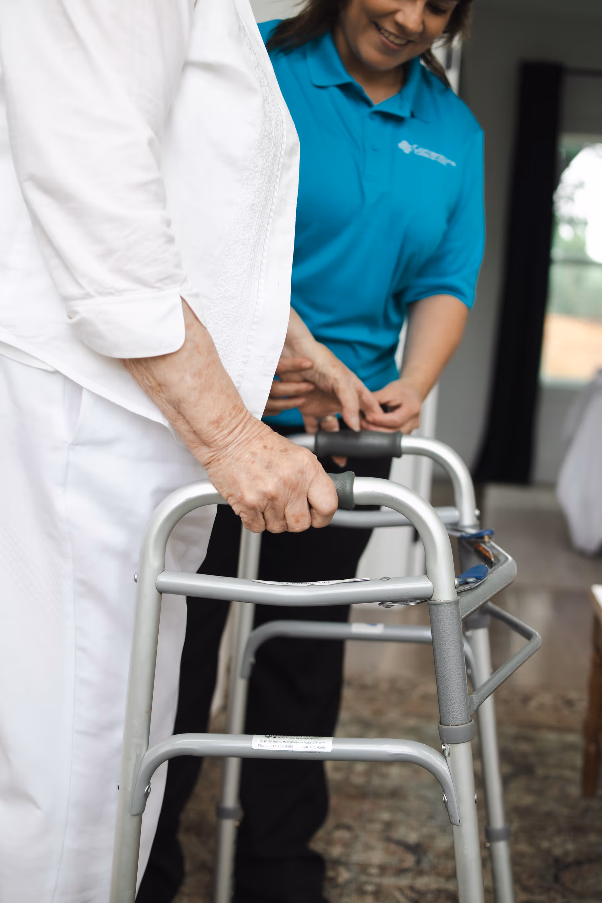 Close-up of hands on a walker, with a caregiver nearby indoors.