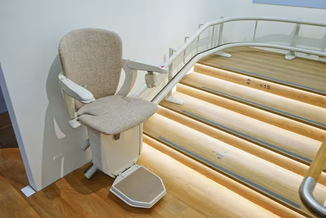Indoor stair lift with beige upholstered seat beside illuminated wooden steps and railing