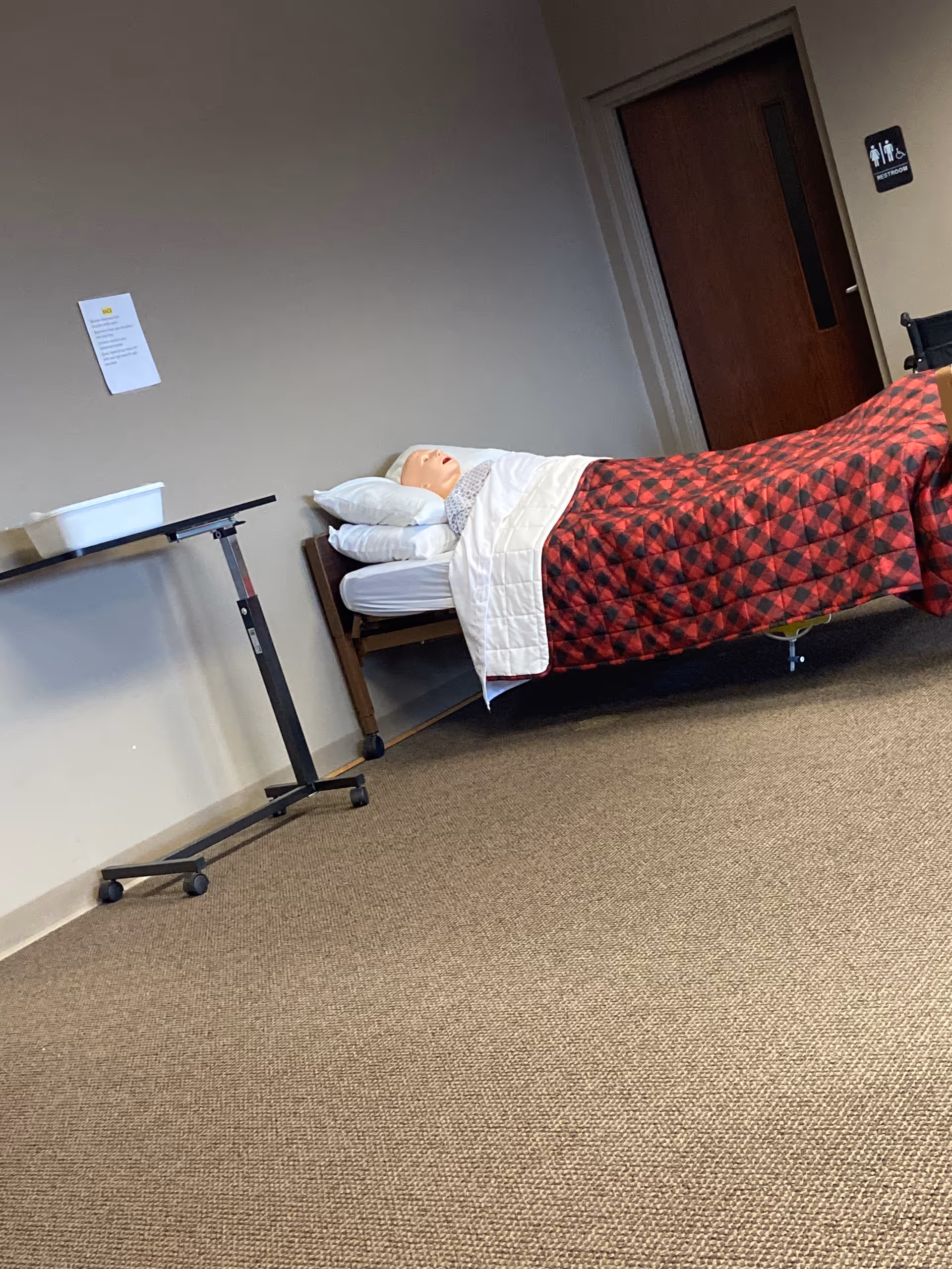 Hospital-style bed with plaid blanket and mannequin head in an empty interior room.