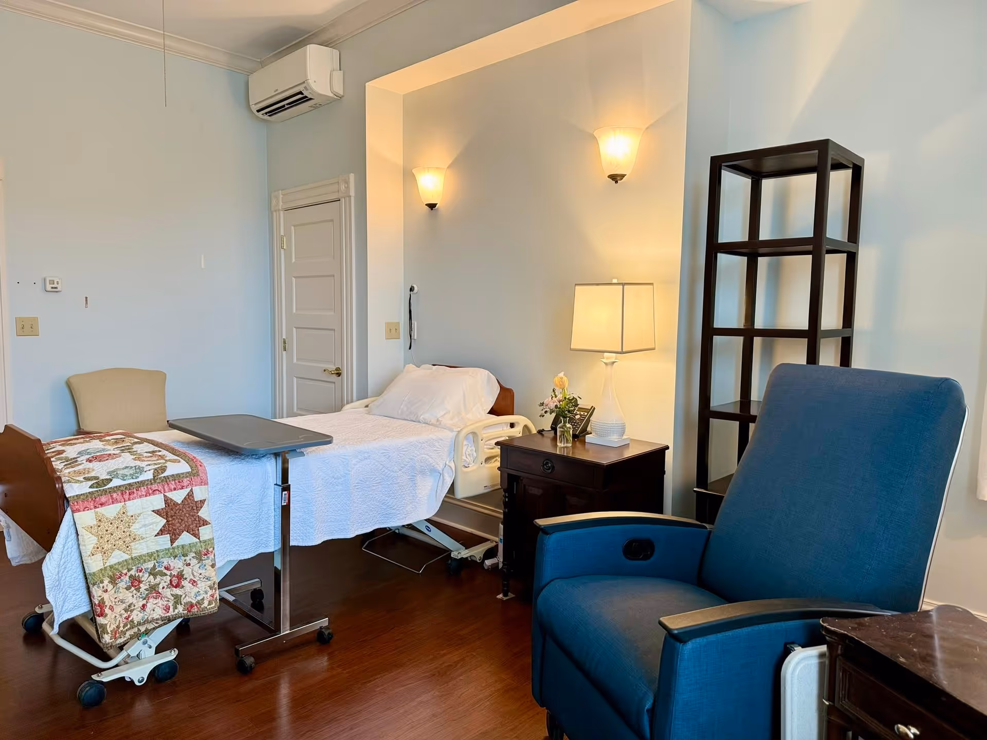 Hospice patient room with a hospital bed, blue chair, quilt, and nightstand.