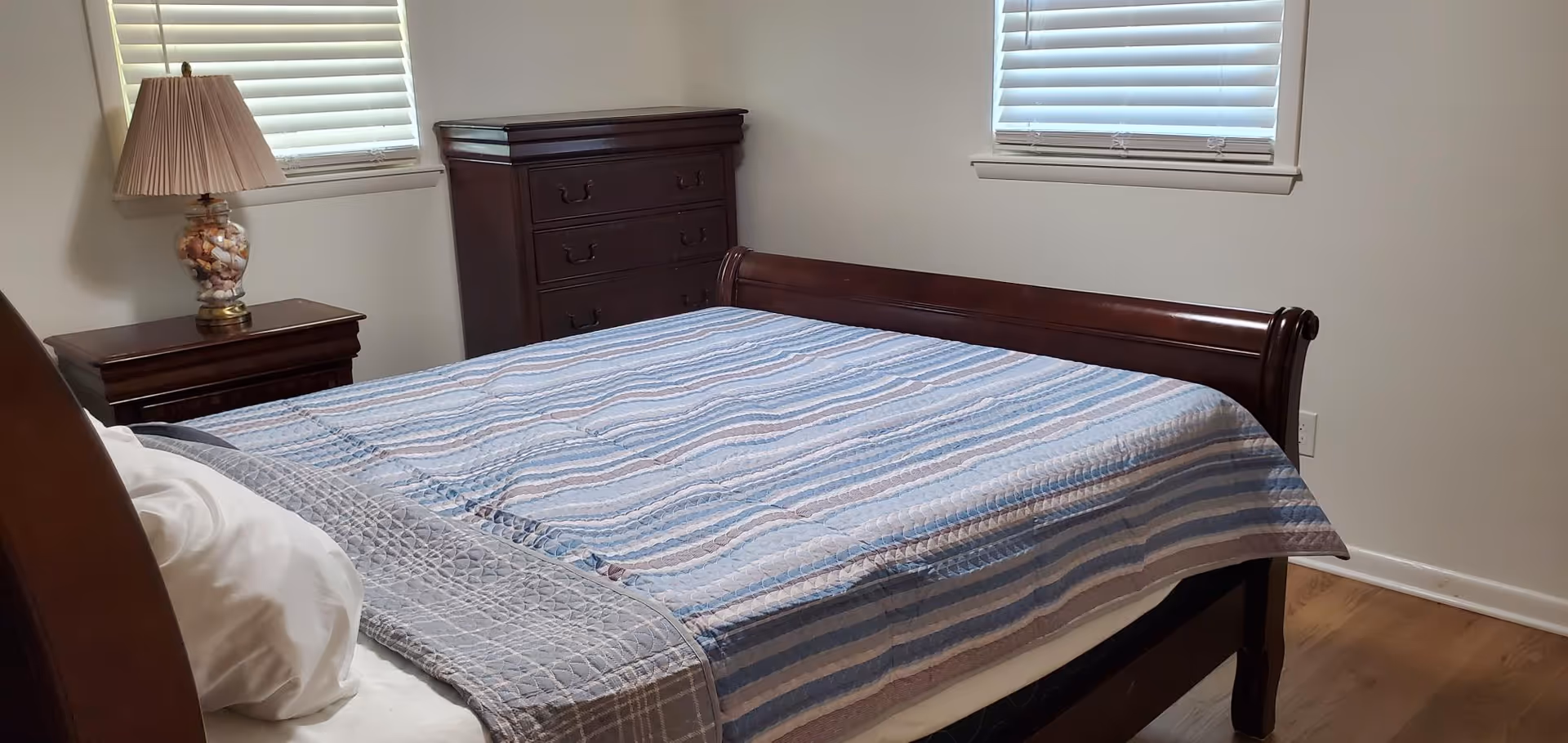 A wooden bed with striped bedding, nightstand, dresser, and two windows.