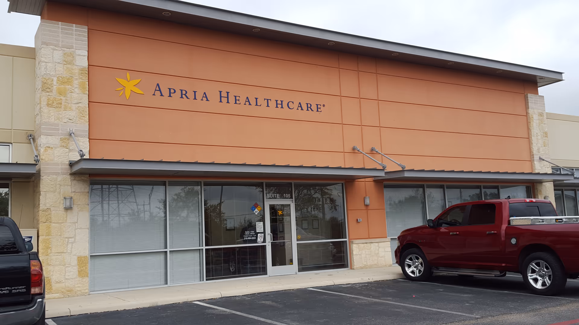 Exterior view of Apria Healthcare building with a glass entrance and a red pickup in parking.
