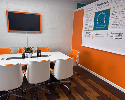 Orange-accented meeting room with a white table, chairs, wall posters, and a mounted screen.