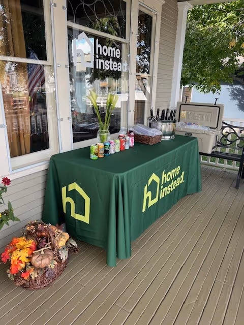 Outdoor porch with a green Home Instead table covered in cloth, beverage cans, and a vase.