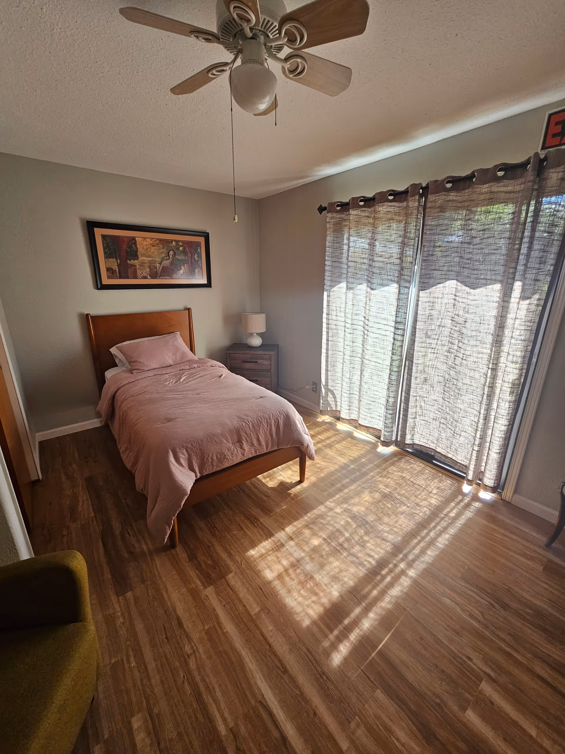 Bedroom with a single bed, pink bedding, nightstand, ceiling fan, and sliding doors with curtains.