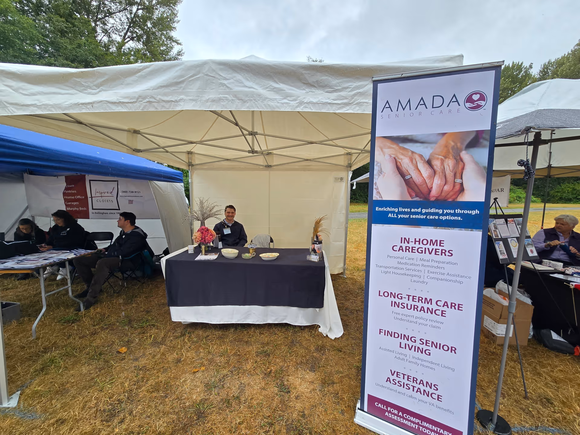 Outdoor Amada Senior Care booth under tents with staff and visitors.