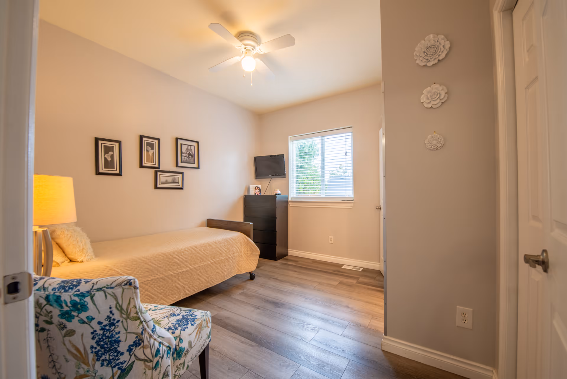 A cozy bedroom featuring a bed, dresser, floral-patterned chair, wall art, and a window.