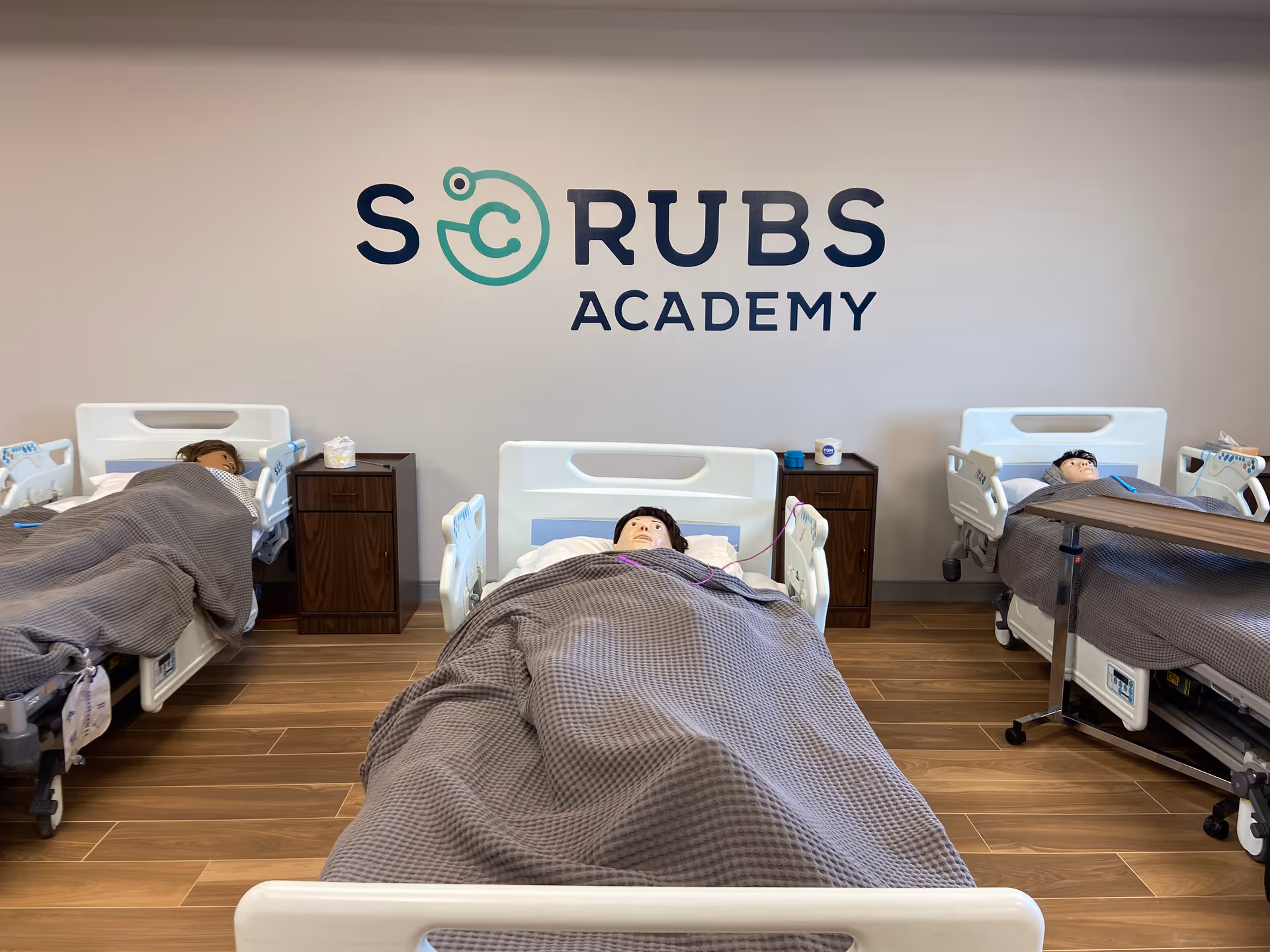 Training room with hospital beds and mannequins under a Scrubs Academy sign.