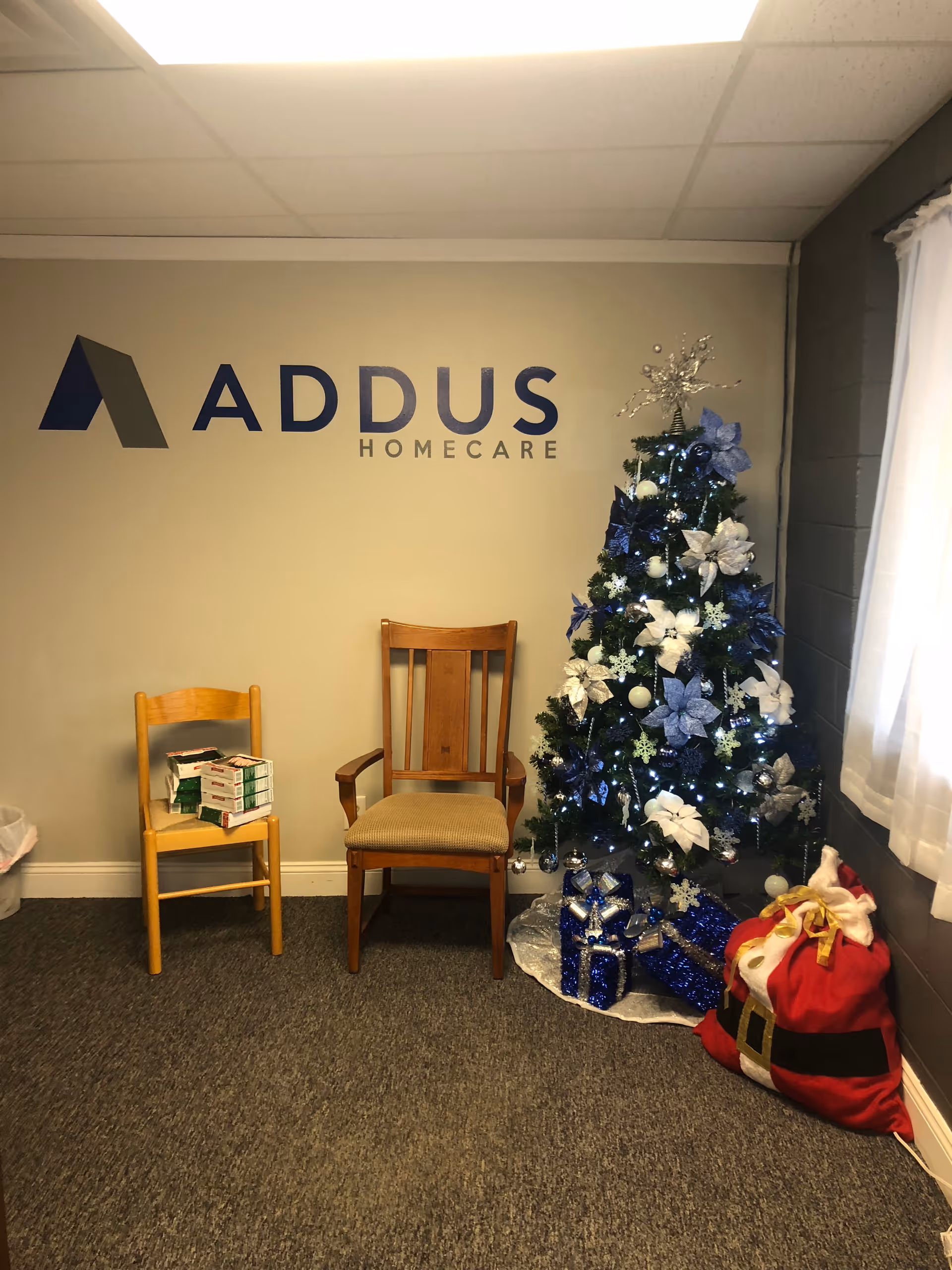 Interior lobby area with the Addus HomeCare logo, Christmas tree, and two wooden chairs.