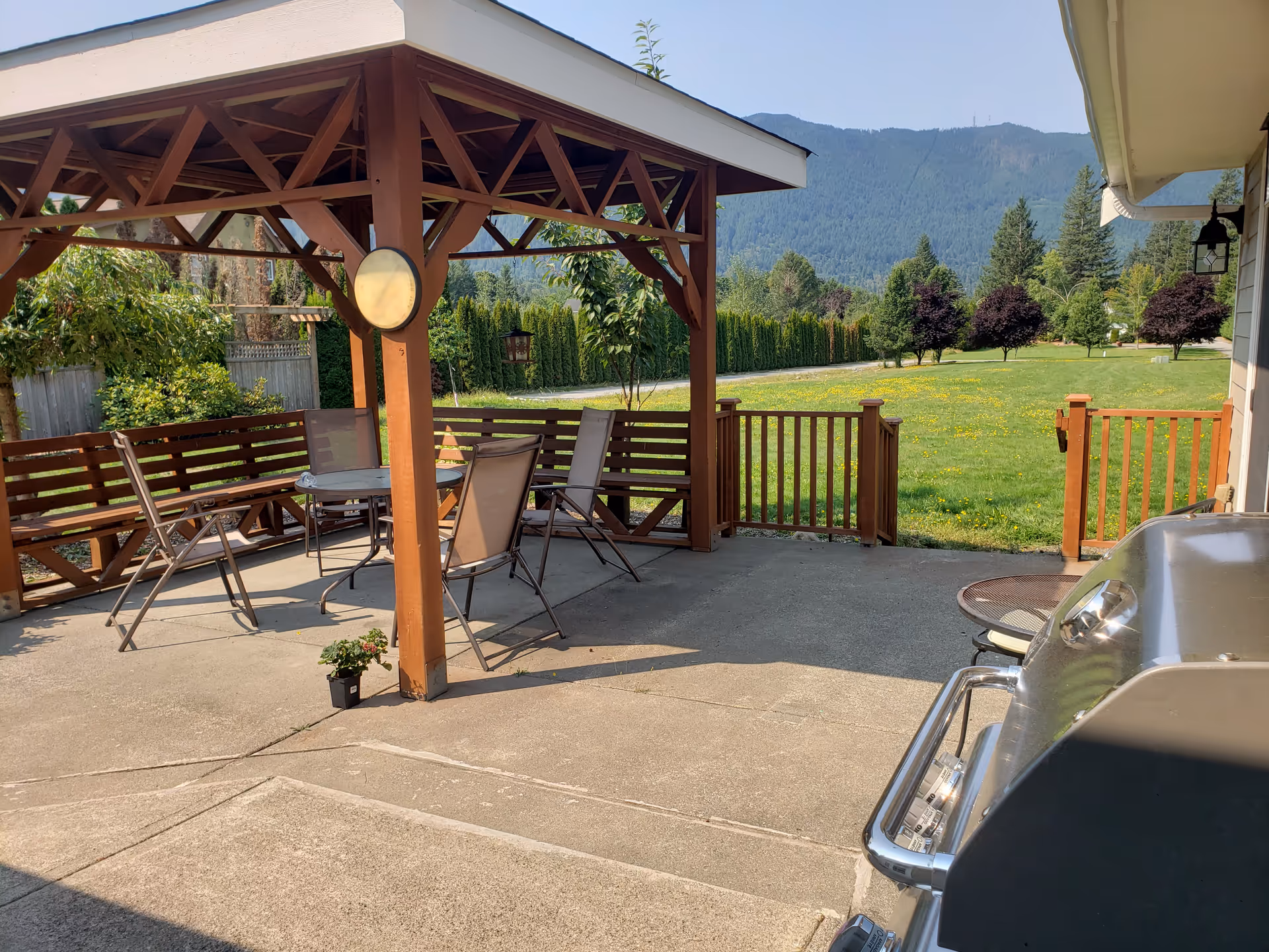 Wooden patio with table and chairs under a pergola overlooking a grassy yard and mountains.