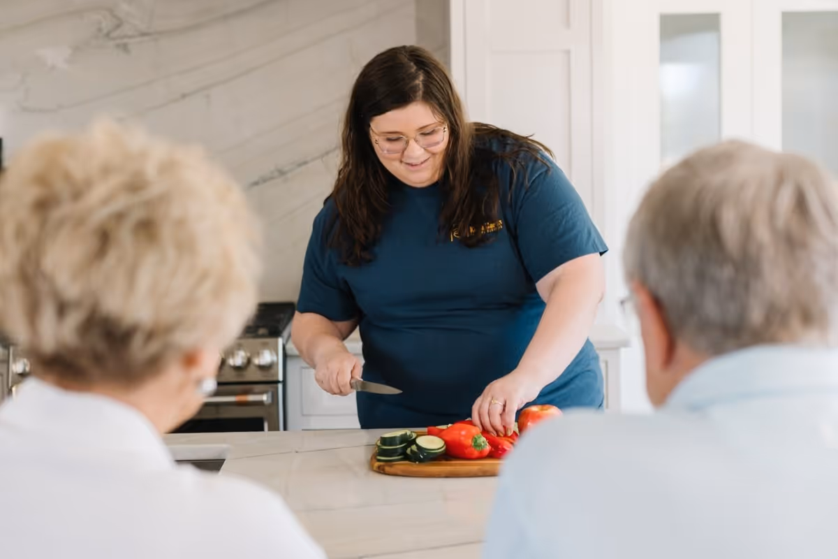 Caregiver chops vegetables at a kitchen island while two residents watch.