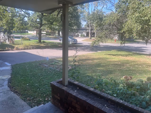 View from a covered porch shows a grassy yard, trees, and a street beyond.