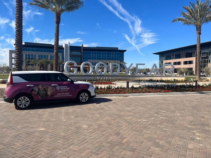 Purple Home Instead van parked before a modern building with Goodyear sign and palm trees.