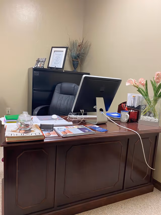 Office desk setup with computer, paperwork, and flowers in a neutral-toned room.