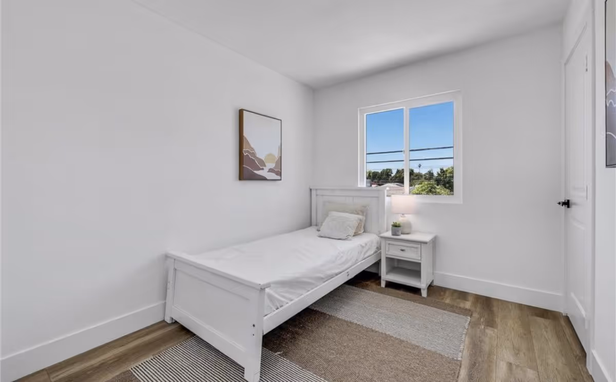 Small white bedroom with a single bed, nightstand, lamp, and a window.