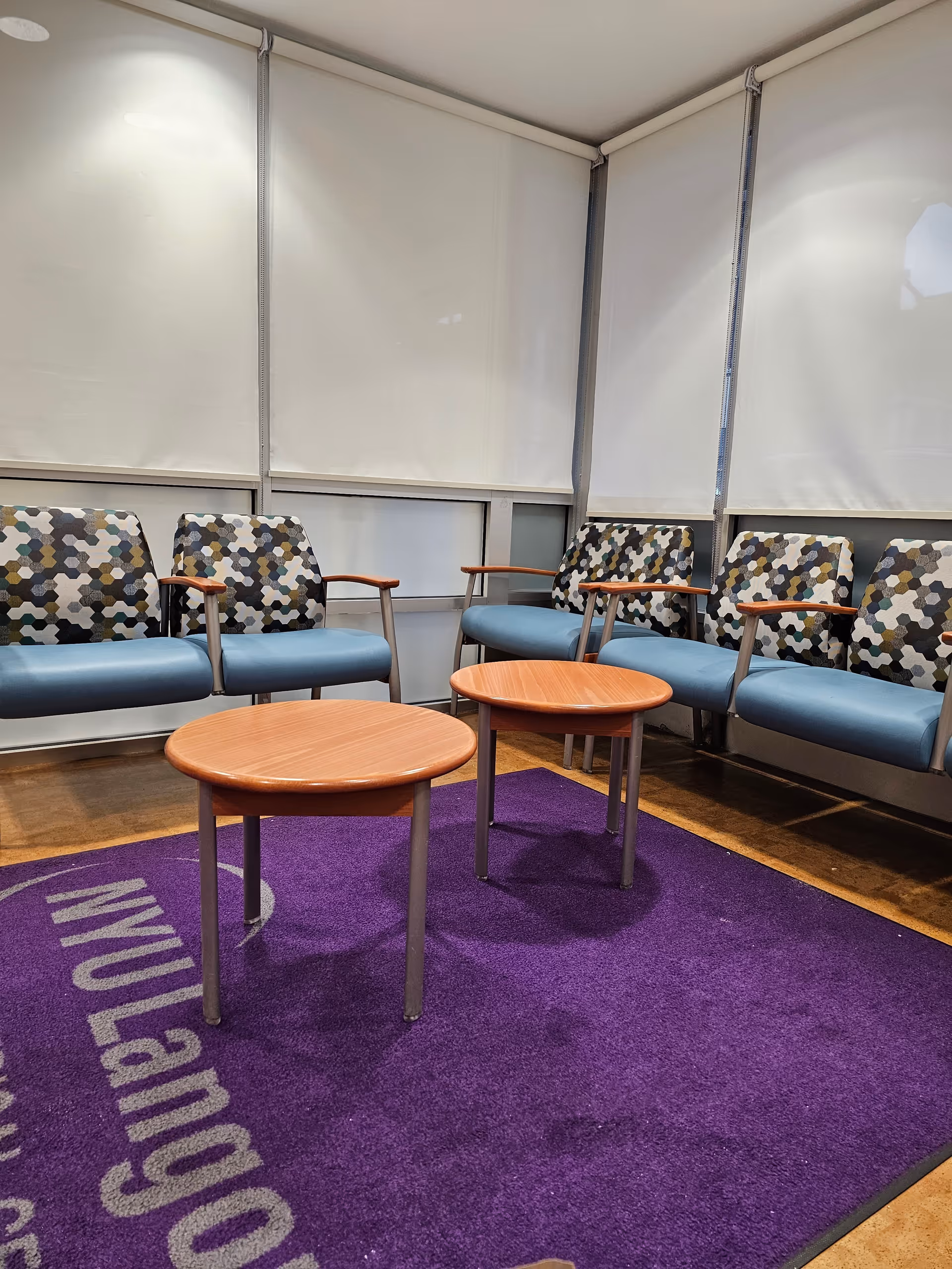 Interior waiting lounge with blue upholstered chairs, wood tables, and a purple NYU Langone rug.