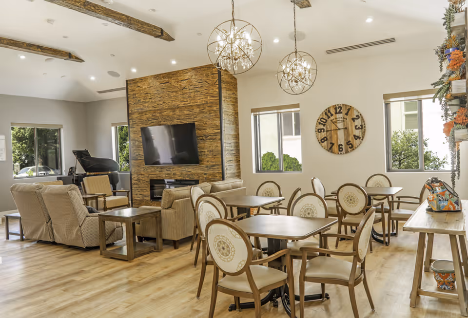 Spacious senior living lounge with sofas, dining tables, fireplace, TV, and large windows.