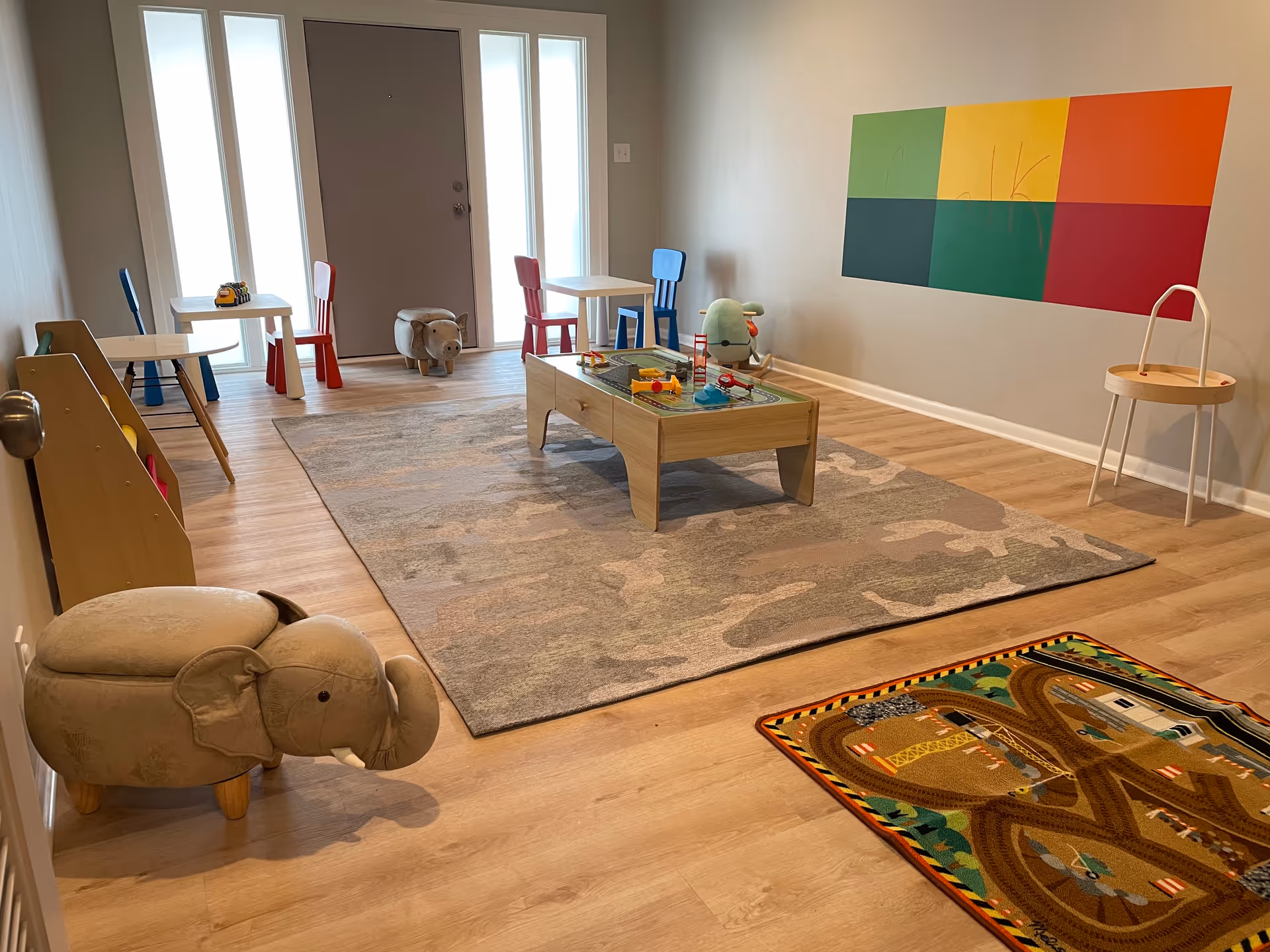 A bright children's playroom with small tables, colorful chairs, toys, and a large rug.
