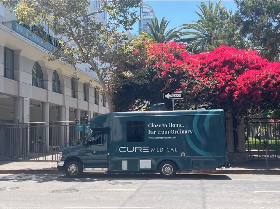 A CURE Medical Care van parked on a city street beside pink flowering bougainvillea.