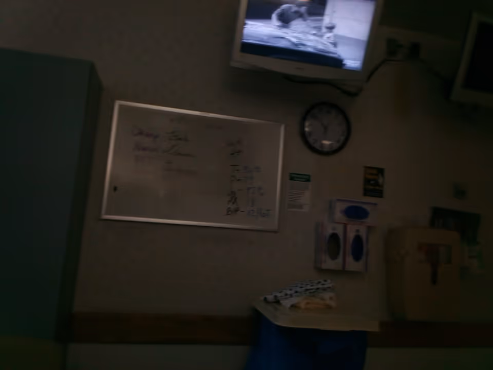 Dim hospital interior featuring a whiteboard, clock, mounted TV, and medical supplies.