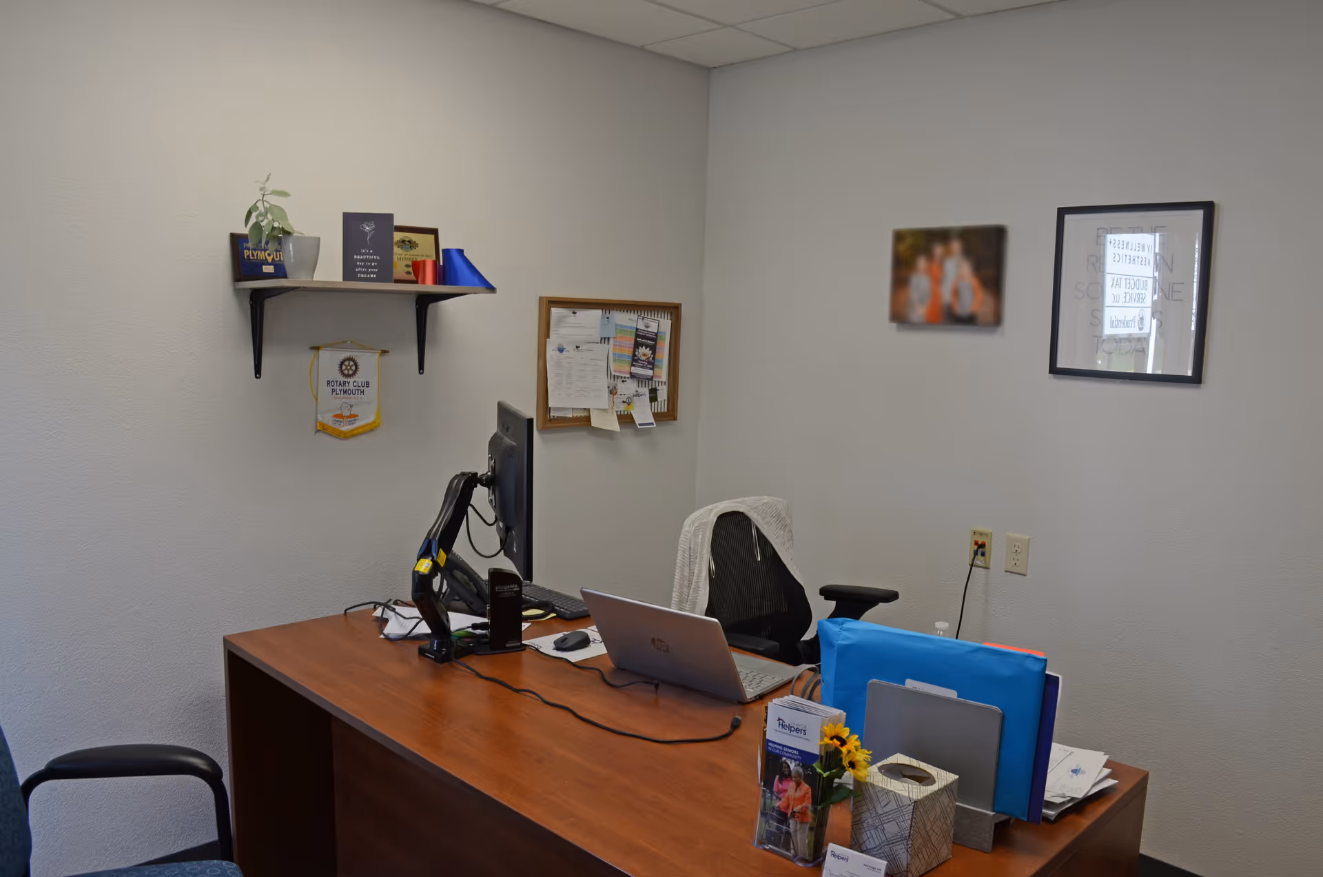 Office desk with computer, monitor, laptop, and bulletin board in a small facility.